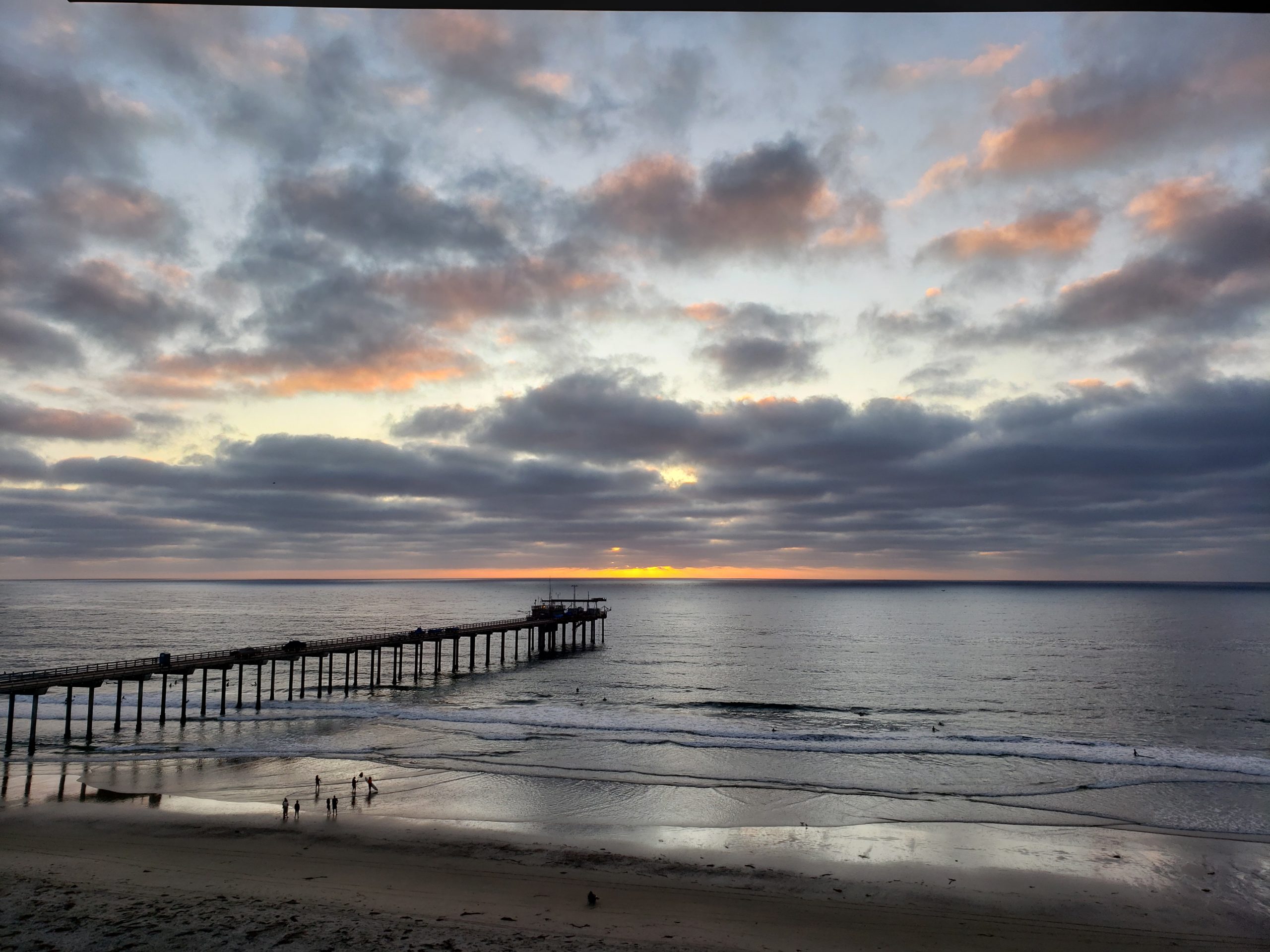 An image showing the Ellen Browning Scripps Pier surrounded by the ocean, a sunset and a cloudy sky. 
